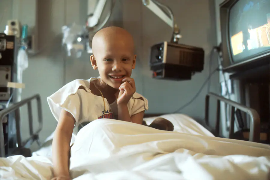 Criança em tratamento de câncer sorrindo sentada no leito do hospital, acompanhada pela cárdio-oncologia pediátrica